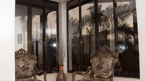 A room with two ornate chairs, a glass-top table, and a large window with a view of palm trees. - UBUNTU PALANCE HOTEL BY RUNATI (Kampala)