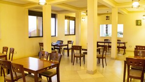 A spacious dining area with multiple wooden tables and chairs, large windows allowing natural light, and modern pendant lighting fixtures. - UBUNTU PALANCE HOTEL BY RUNATI (Kampala)