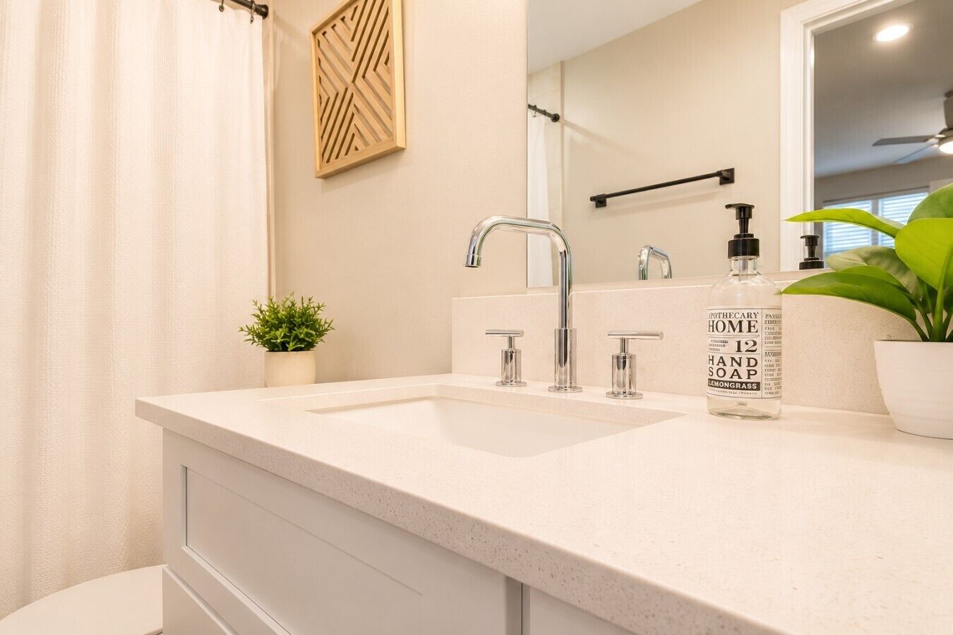 A bathroom with a white countertop, two sinks, a mirror, a shower curtain, a potted plant, and a bottle of soap.