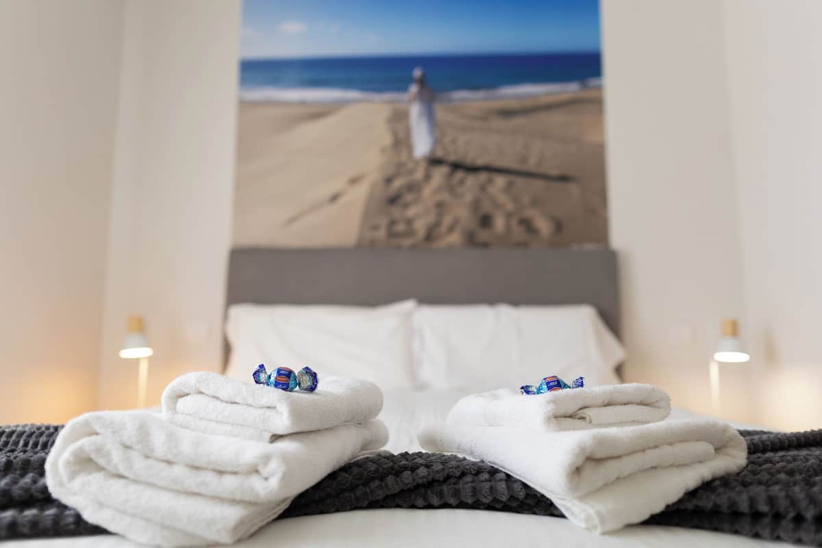 A bed with white towels folded at the foot, a gray knitted blanket, and a poster of a beach scene with a person walking on the sand.