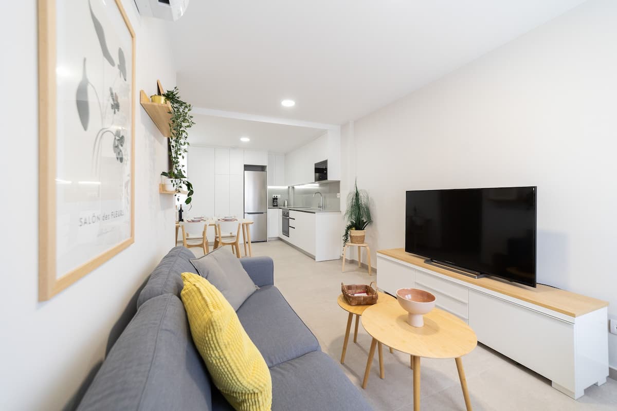 A modern living room with a grey sofa, a yellow throw pillow, a wooden coffee table, and a flat-screen TV on a white cabinet.