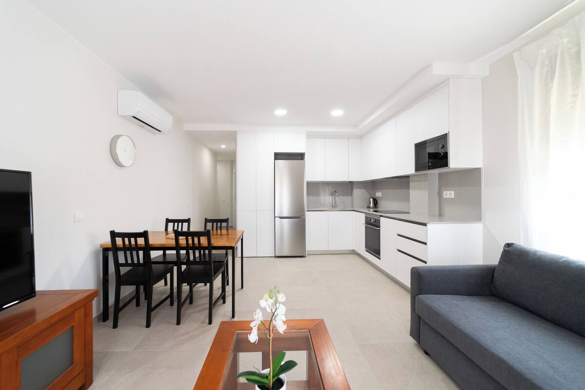 A modern living room with a grey sofa, wooden dining table, and chairs. There is a TV on a wooden stand, a small table with a plant, and a kitchen area with white cabinets and appliances.