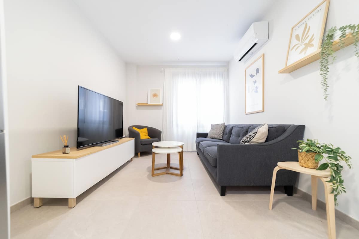 A modern living room with a flat-screen TV, a grey sofa, a small round table, and a potted plant.