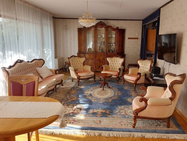 Ein traditionell eingerichtetes Wohnzimmer mit einem großen Holzschrank, mehreren Polstersesseln, einem gemusterten Teppich und einem Fernseher.