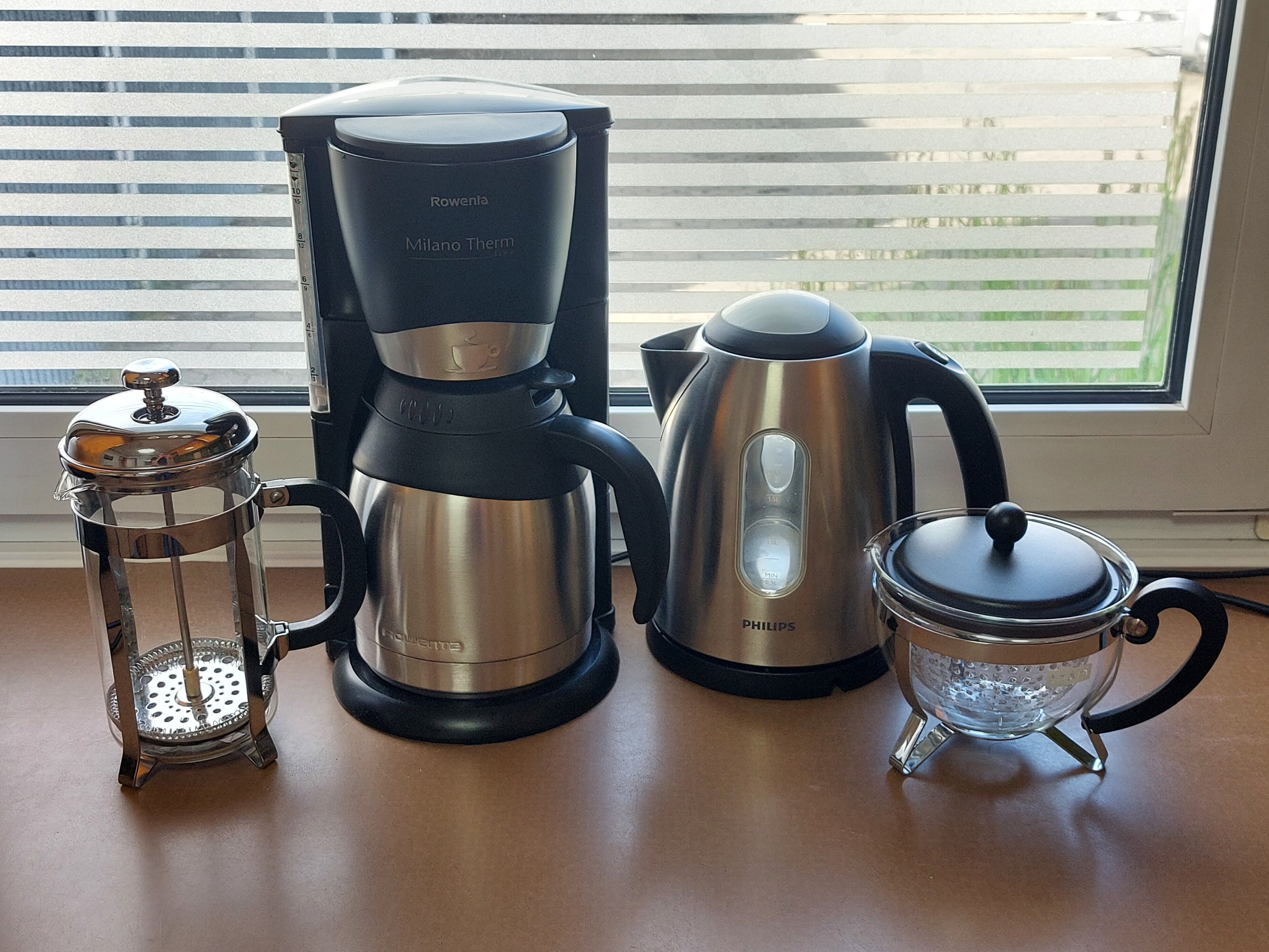 Uma cafeteira, um bule e uma prensa francesa sobre uma mesa perto de uma janela.