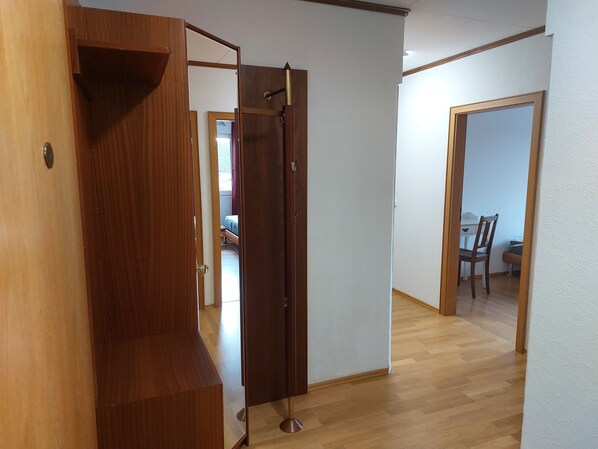 A hallway with wooden flooring, a door with a brass handle, and a visible bed in the background.