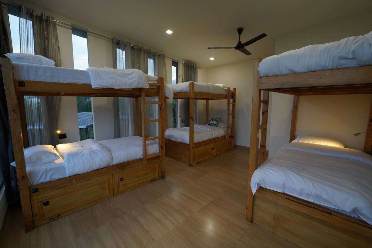 Una habitación con tres literas, suelo de madera, ventilador de techo y ventanas grandes con cortinas.