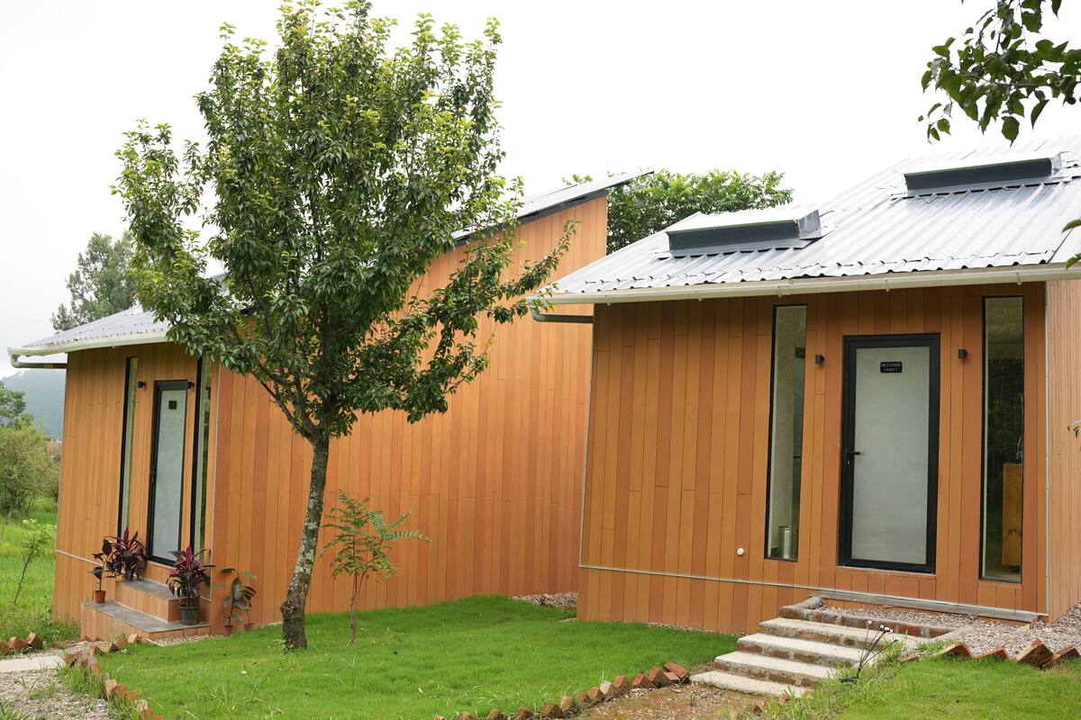Un edificio moderno de madera con techo metálico, un árbol pequeño al frente y un camino pavimentado que lleva a la entrada.