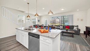 A spacious kitchen with white cabinets, a large island, and a dishwasher. There is a dining area with a gray sofa and a coffee table. - Poolside Delight. Whimsical Themed Rooms and Ps5! (Davenport)