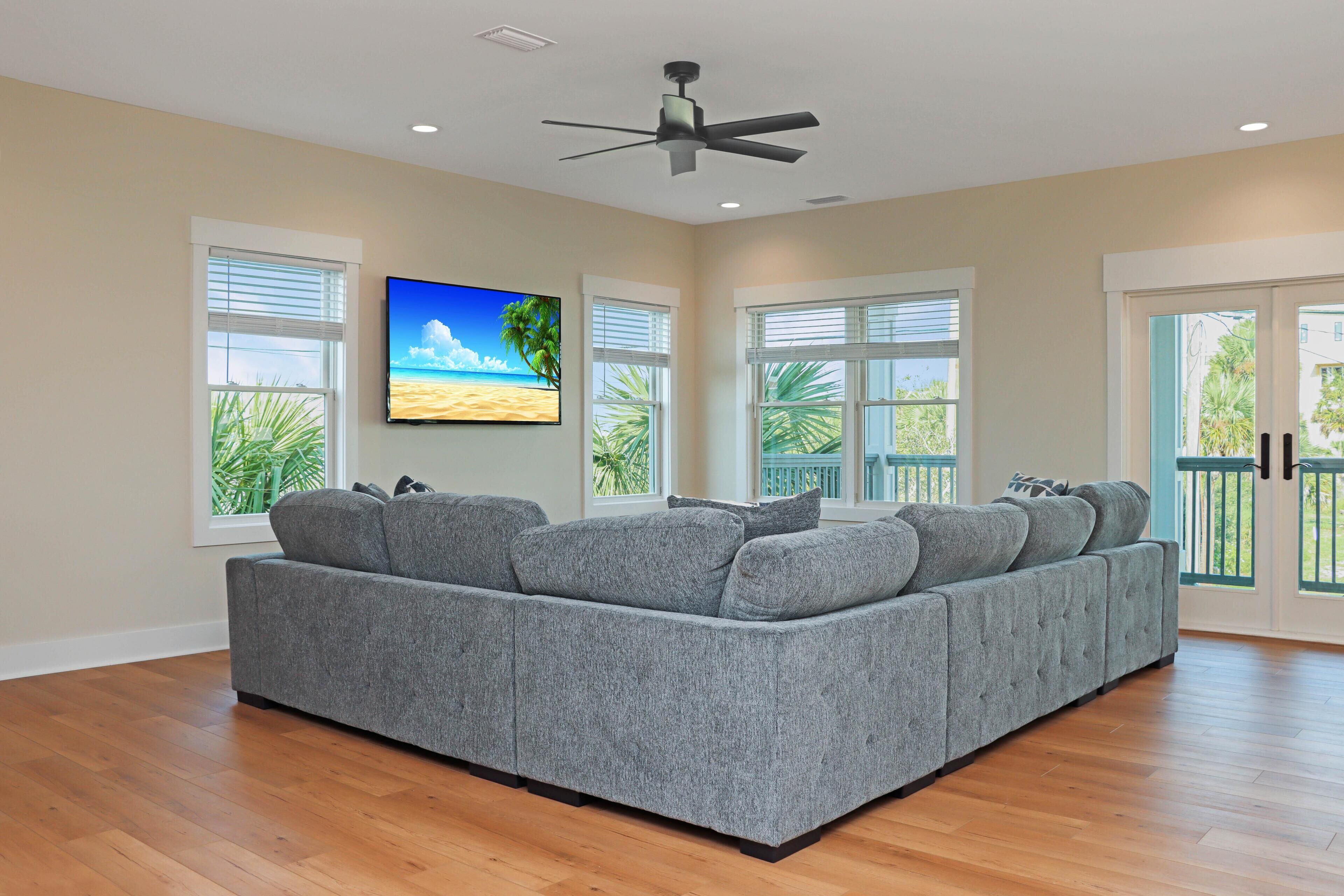 A spacious living room with a grey sectional sofa, large windows with blinds, a ceiling fan, and a flat-screen TV on the wall.