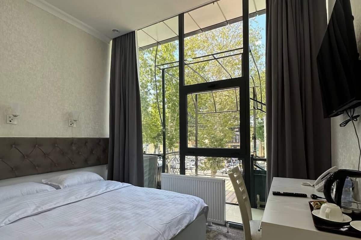 A hotel room with a large bed, a desk with a telephone and a kettle, and a view of trees through large windows.