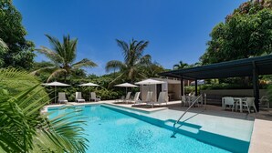 En swimmingpool med liggestole og parasoller, omgivet af palmer og frodig vegetation.