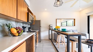 Et moderne køkken med træskabe, et rustfrit stålkøleskab og en skål med frugt på bordet. Der er et lille spiseområde med et bord og barstole.