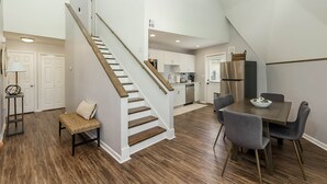 A spacious living area with a wooden staircase, a dining table with chairs, and a kitchen in the background.