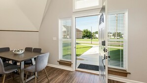A dining area with a table and chairs, a window with blinds, and a view of a green lawn and a building outside.