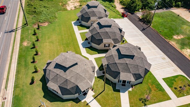 A residential area with four houses featuring unique geodesic dome roofs, arranged in a row with a paved driveway and a road nearby.