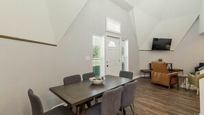 A dining area with a wooden table and grey chairs, a small side table with a plant, and a television mounted on the wall.