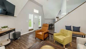 Een ruime woonkamer met hoge plafonds, een wandtelevisie, een eethoek met tafel en stoelen, en een trap naar een ander niveau.