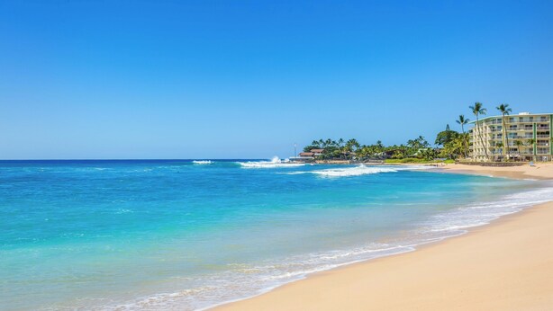 Una playa con agua azul cristalina, orilla arenosa y un edificio de resort con palmeras.