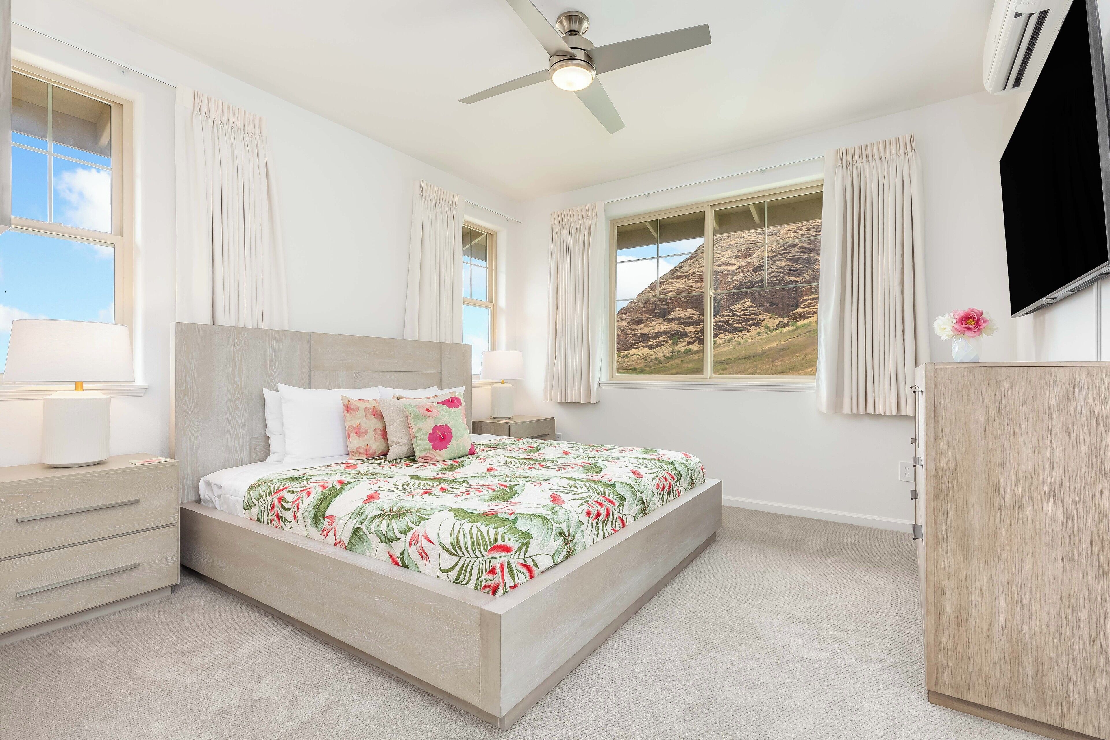A bedroom with a bed, nightstands, a ceiling fan, a TV, and large windows with curtains.