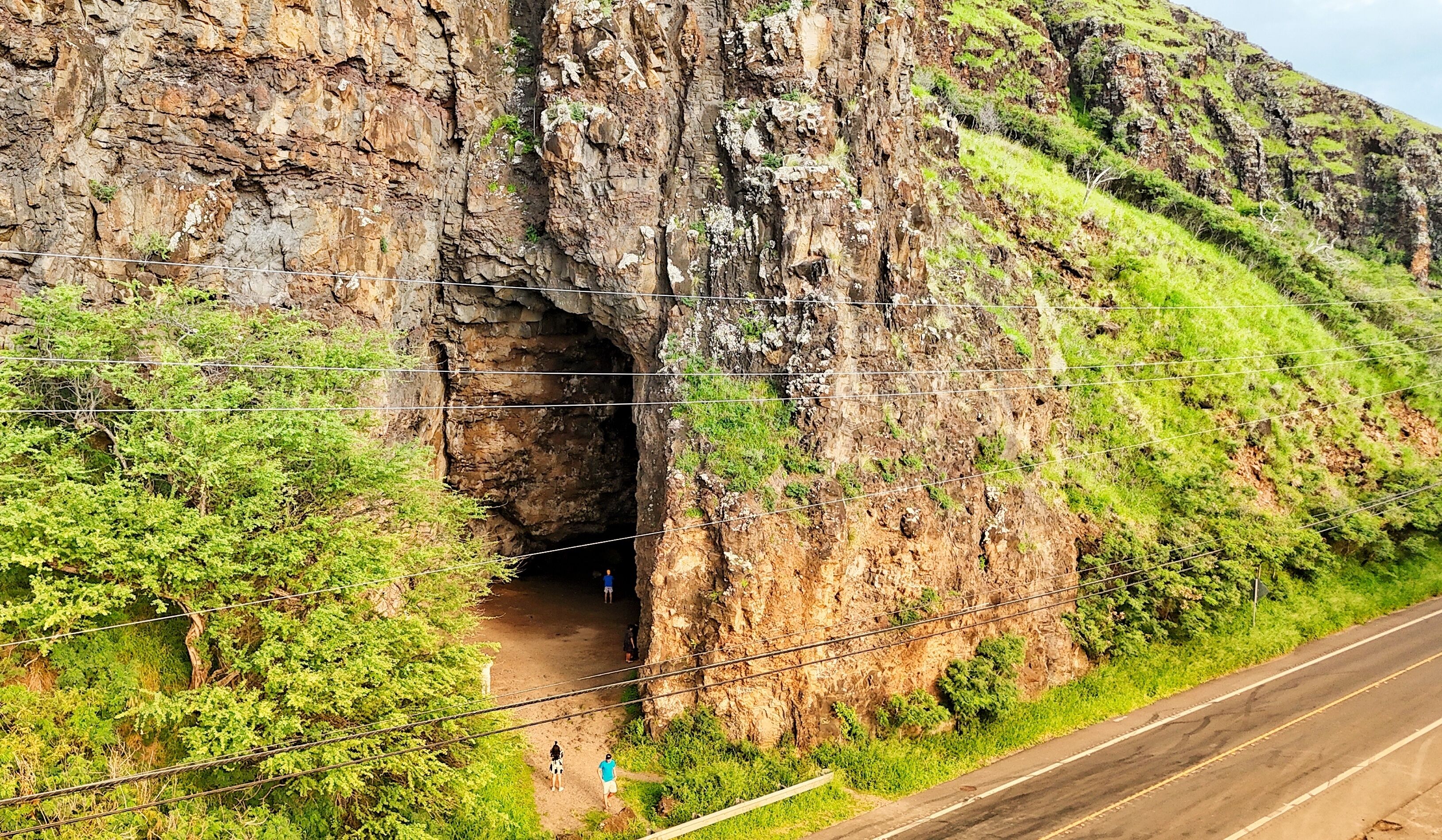 A rocky cliff with a cave entrance, a road beside it, and some greenery.