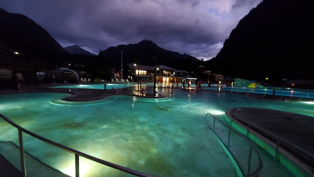 Une piscine à la forme incurvée, entourée d’une balustrade, et un bâtiment illuminé en arrière-plan.