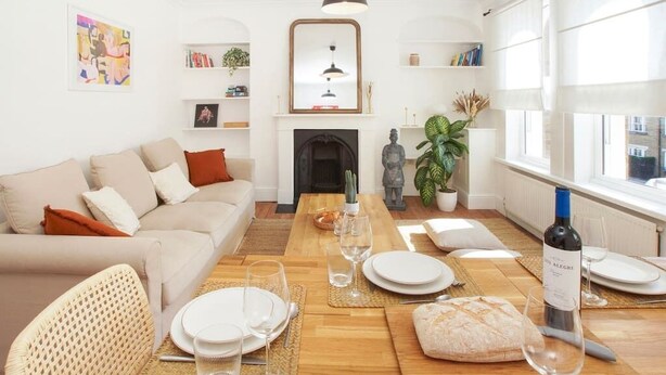 A living room with a sofa, a dining table set for a meal, a fireplace, and a large window. - Spacious and stylish flat in the vibrant neighbourhood of Mile End, East London! (Greater London)