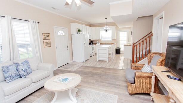 A spacious living room with a white sofa, a round coffee table, and wicker chairs. A kitchen is visible in the background with white cabinets and a wooden staircase. - Beautiful Quaint Home Half a Block from the Beach! W/Gulf Views! (Panama City Beach)