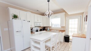A kitchen with white cabinets, a white refrigerator, a white island with bar stools, a microwave, a coffee maker, and a chandelier. - Beautiful Quaint Home Half a Block from the Beach! W/Gulf Views! (Panama City Beach)