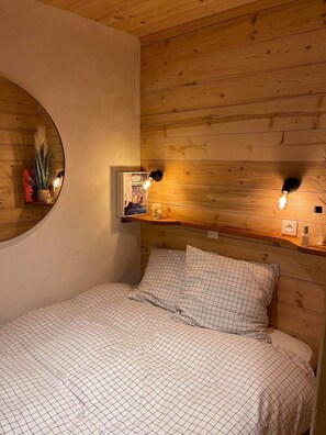 A bedroom with a wooden ceiling, a round mirror, a bedside table with a lamp, and a bed with a checkered duvet.