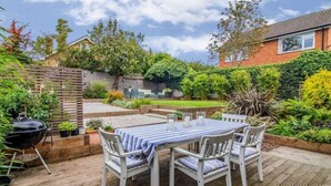 A patio with a table and chairs, surrounded by a garden and a brick house.