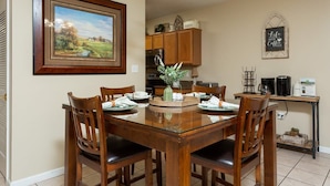A dining area with a wooden table and chairs, a framed landscape painting, and a kitchen in the background. - Riverfront Condo Pigeon Forge (Pigeon Forge)
