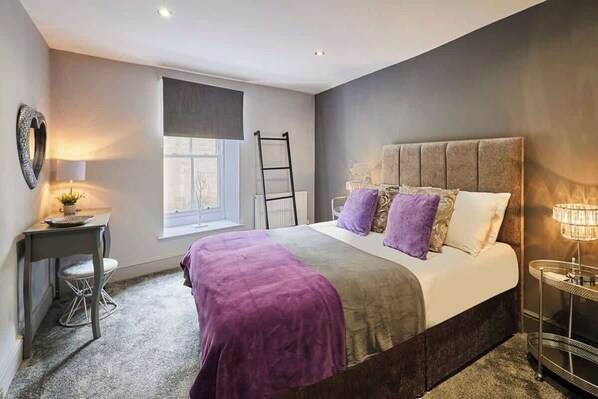 A bedroom with a bed, a desk, a chair, a window, and a nightstand with a lamp.