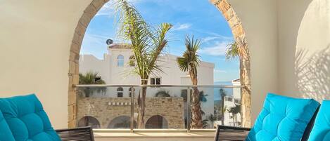A balcony with two blue cushioned chairs and a glass-top table, overlooking a building with a stone archway and palm trees.