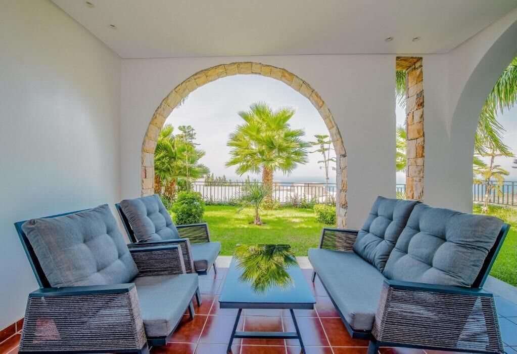 A patio with two cushioned chairs and a table, an archway leading to a garden with palm trees, and a view of the sea.