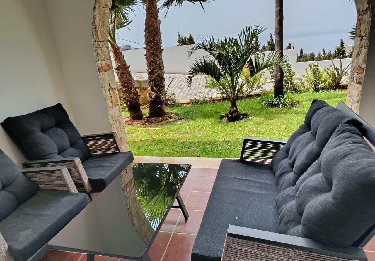 Ein moderner Außenbereich mit grauen Sofas und einem Glastisch, mit Blick auf einen gepflegten Garten mit Palmen und einer weißen Wand im Hintergrund.