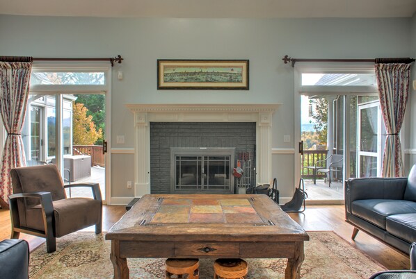 A living room with a fireplace, a large wooden table, and a sofa. There are two chairs and a rug on the floor.