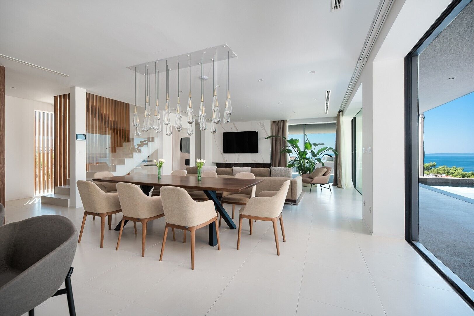 A modern dining area with a large table and chairs, a staircase, and a view of the sea.