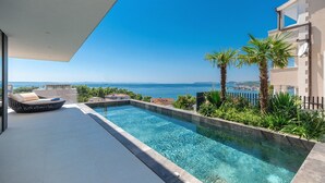 Un área de piscina moderna con vista despejada al mar, una silla de descanso y abundante vegetación.