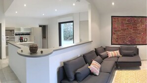 A modern living area with a grey sofa set, a kitchen with white cabinets, and a large window providing natural light.