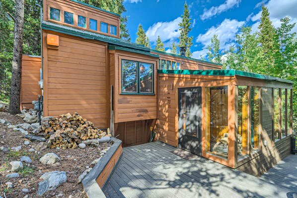Une cabane en bois dotée d’une terrasse, de grandes fenêtres et d’une cheminée en pierre.