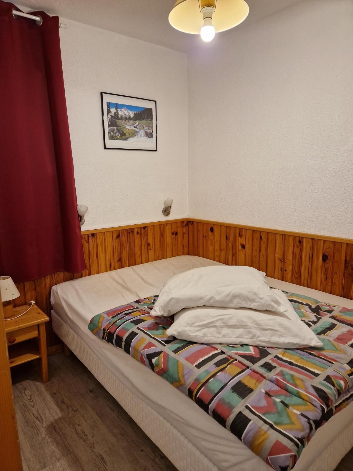 A single bed with a colorful blanket and white pillows, a wooden nightstand with a lamp, a red curtain, and a framed picture on the wall.