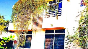 Un edificio blanco con balcón, rodeado de una cerca enrejada blanca y diversas plantas.