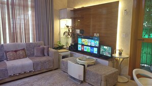 Una sala de estar con un sofá, una mesa de centro, un televisor mostrando varios canales y una pequeña mesa con una planta.