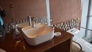 Une salle de bain avec un lavabo blanc, un miroir et un mur en pierre. Un flacon de savon liquide est posé sur le comptoir.