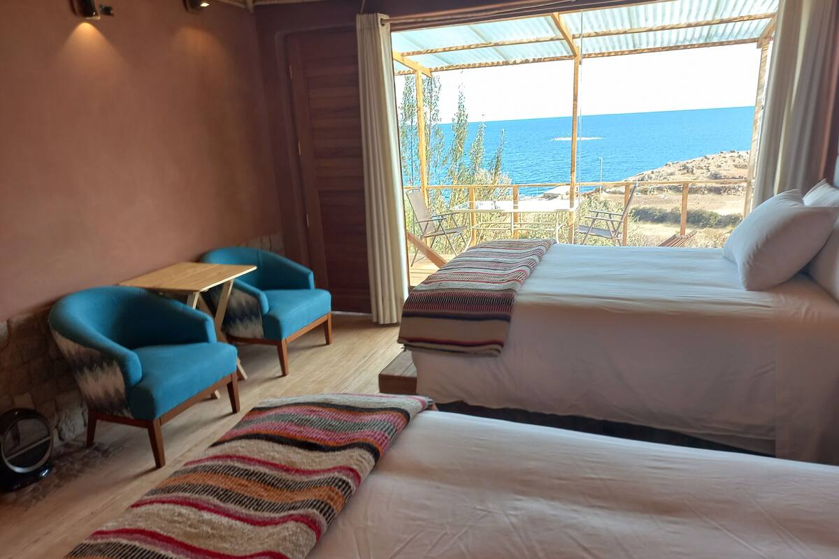 A room with two beds, a small table, and two chairs. There is a view of the sea through a window.