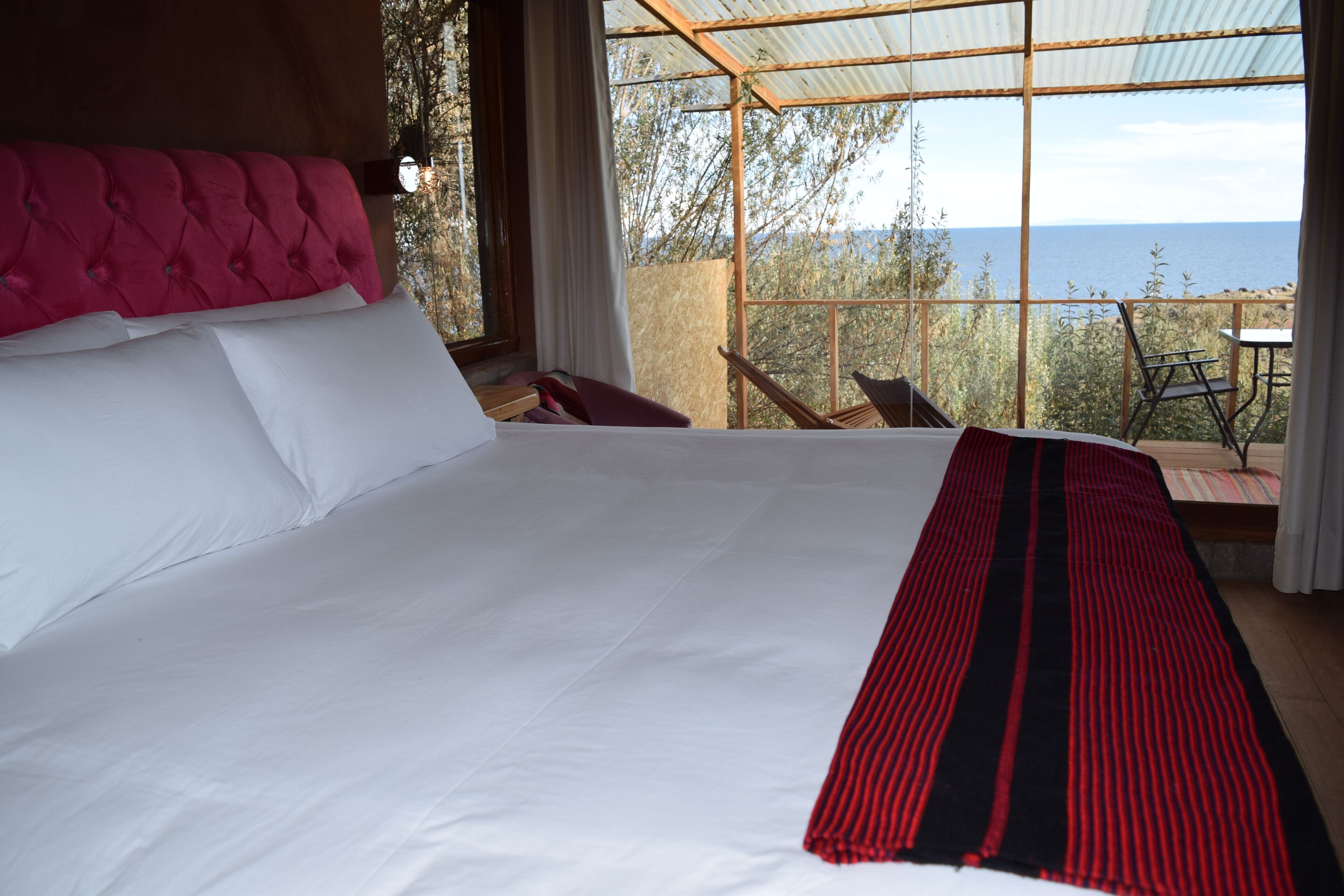 Un grand lit avec du linge de lit blanc et une couverture à rayures rouges et noires, une tête de lit rose à motifs capitonnés, et une vue sur la mer par une grande fenêtre.