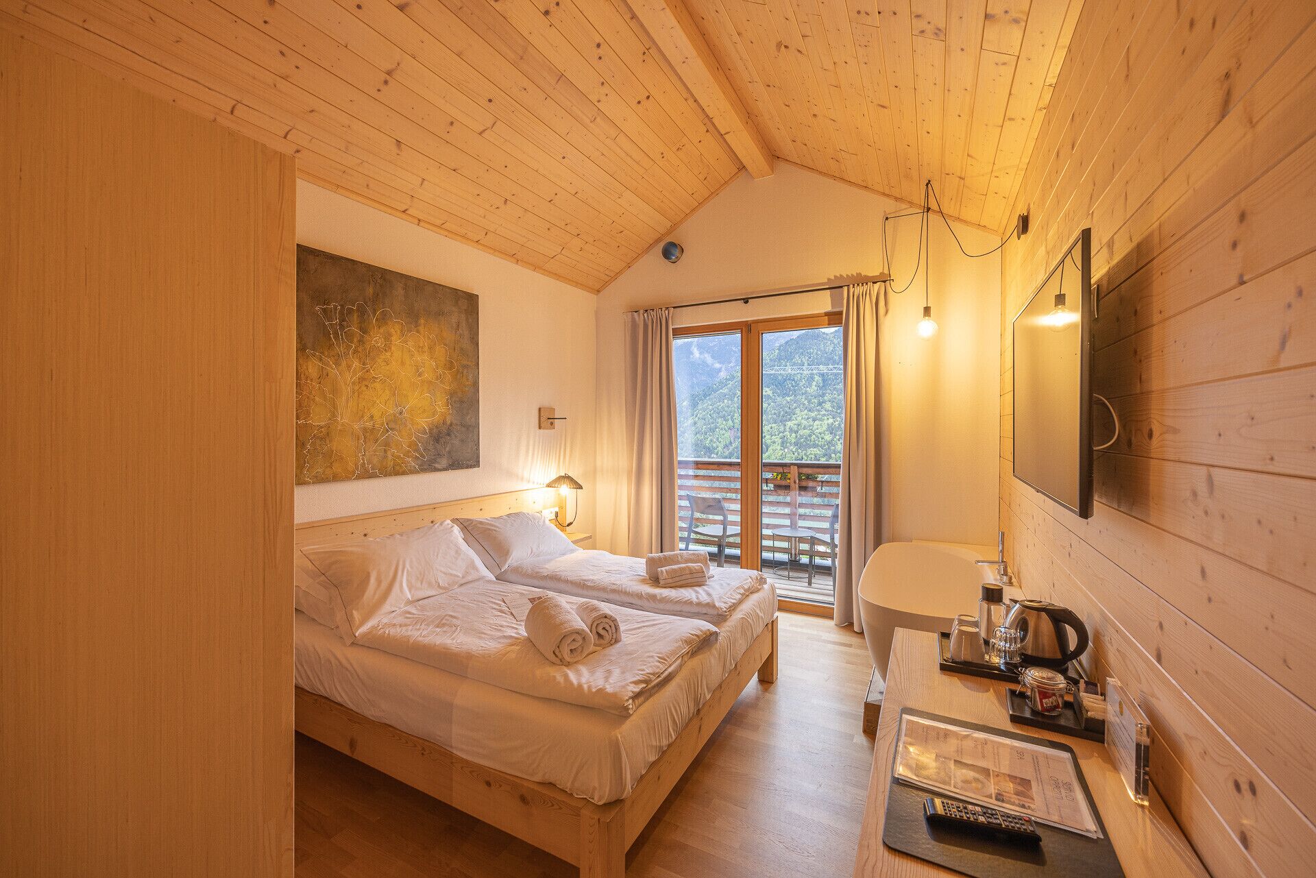 Una habitación acogedora de madera con dos camas, un balcón con vistas a la montaña, un escritorio con hervidor y un cuadro en la pared.
