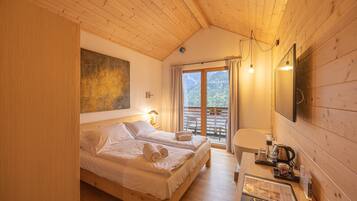 A cozy wooden room with two beds, a balcony with a mountain view, a desk with a kettle, and a painting on the wall.