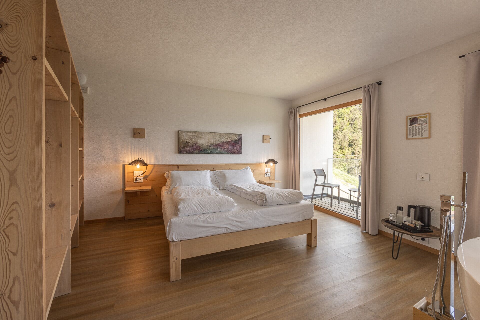 Une chambre à coucher avec un lit en bois, des lampes de chevet, un tableau, une fenêtre donnant sur un paysage verdoyant et une salle de bain avec douche.